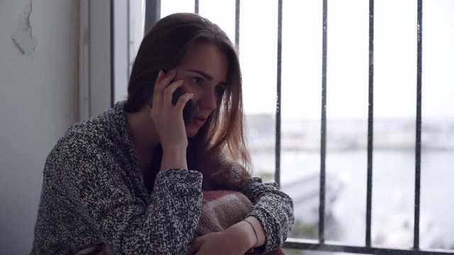 Young attractive woman is talking over the mobile phone laughing at the end. Lonely depressed woman sitting by the window.