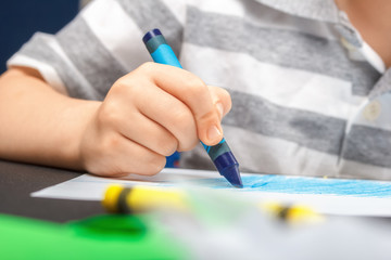 Drawing on a white sheet of paper with colored wax pencils. The concept of children's creativity and hobbies. The child draws a blue pencil on white paper. Baby hands closeup