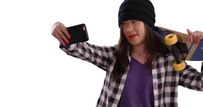 Cool millennial skater taking selfie with smartphone camera and holding skateboard on white background, Portrait of young Asian woman using technology to take picture for copy space, 4k