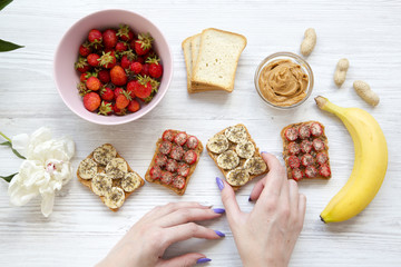 Young girl takes vegan toast with fruits, seeds, peanut butter over white wooden background, top view. Healthy breakfast with ingredients, dieting concept. From above.