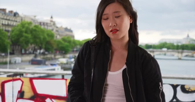 Chinese woman in black bomber jacket on bridge over the River Seine with hair blowing in wind, Asian millennial tourist waits on sidewalk with pockets in Paris, 4k