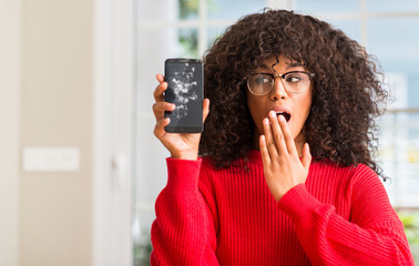 African american woman holding broken smartphone cover mouth with hand shocked with shame for...