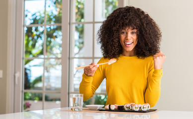 African american woman eating sushi using chopsticks at home screaming proud and celebrating victory and success very excited, cheering emotion