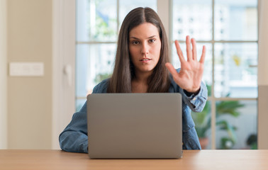 Young woman using laptop at home with open hand doing stop sign with serious and confident...