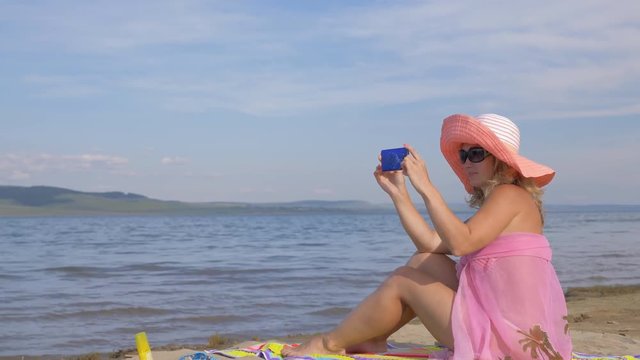 Woman with cell phone on the beach. She rewrites by phone, looks at photos, takes a photo of herself, selfie. The girl is taking pictures of the sea on the phone.