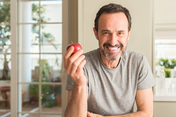 Middle age man eating healthy red apple with a happy face standing and smiling with a confident...