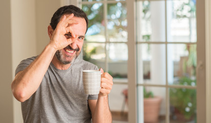 Middle age man drinking coffee in a cup with happy face smiling doing ok sign with hand on eye looking through fingers
