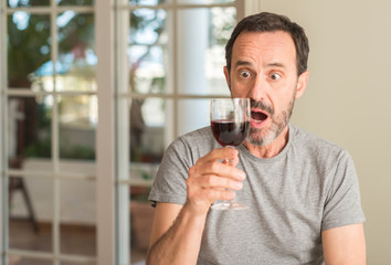 Middle age man drinking a glass of wine scared in shock with a surprise face, afraid and excited...