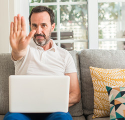 Middle age man using laptop at sofa with open hand doing stop sign with serious and confident expression, defense gesture