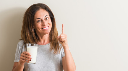 Middle age woman drinking a glass of fresh milk surprised with an idea or question pointing finger with happy face, number one