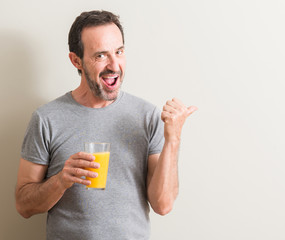 Senior man drinking orange juice in a glass pointing with hand and finger up with happy face smiling