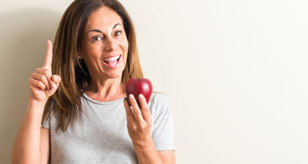 Middle age woman holding a green apple surprised with an idea or question pointing finger with happy face, number one