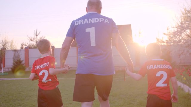 Backshot of grandfather is handling his grandsons, wearing football uniform, with soccerball