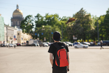 Fototapeta premium Back view of young stylish man tourist walking at Palace square near Saint Isaac's Cathedral in Saint-Petersburg. He wearing in black jeans, black T-shirt, black cap and red backpack