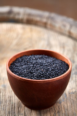 Black sesame seeds in a clay bowl on wooden barrel background, close-up, top view, vertical.