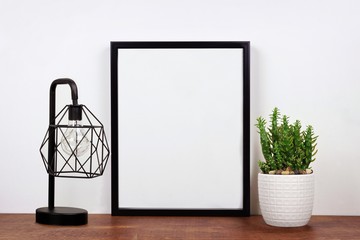 Mock up black frame, cactus plant and industrial style lamp on a shelf or desk. Wood shelf and white wall. Portrait frame orientation.