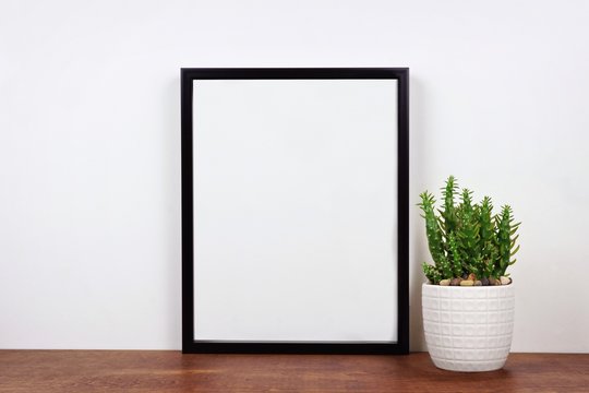 Mock up black frame with cactus plant on a shelf or desk. Wood shelf and white wall. Portrait frame orientation.