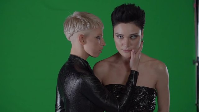 Two girlfriends are standing in background of chroma key green-screen. Blonde girl is seducing brunette, touching her by hand