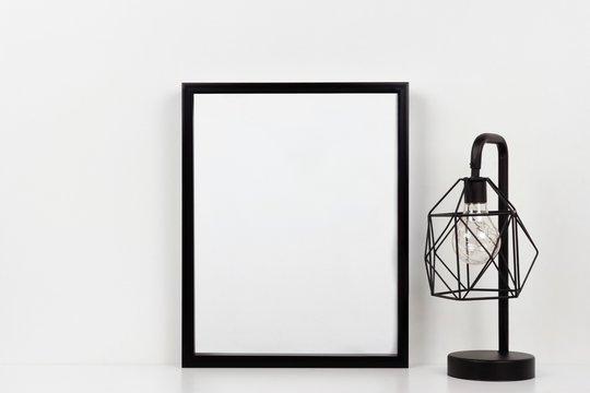 Mock up black frame and industrial style lamp on a shelf or desk. White shelf and wall. Portrait frame orientation.