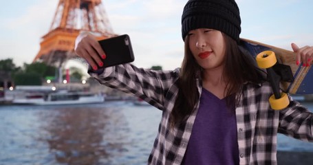 Cool millennial skater taking selfie with smartphone camera and holding skateboard in Paris France, Portrait of young Asian woman using technology to take picture in front Eiffel Tower, 4k - Powered by Adobe
