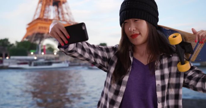 Cool millennial skater taking selfie with smartphone camera and holding skateboard in Paris France, Portrait of young Asian woman using technology to take picture in front Eiffel Tower, 4k