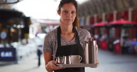 Unhappy waitress holding tray with coffee and tea outside, Portrait of serious attractive woman wearing apron and carrying tray to serve beverages, 4k