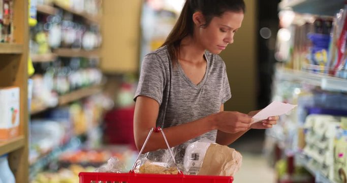 Pretty young woman at grocery store checking her shopping list, Young wife or mother customer carrying basket looking for item in store aisle, 4k