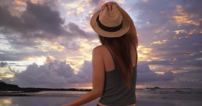 Cute millennial girl on vacation twirling on the beach with fedora, Joyful woman in striped tank top looking out at ocean and dancing, 4k