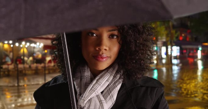 Close up of African American woman standing under black umbrella on rainy night in paris, Millennial tourist in rain coat waiting on street in the rain, 4k