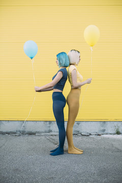 Two female friends sitting in front of a yellow wall and holding balloons