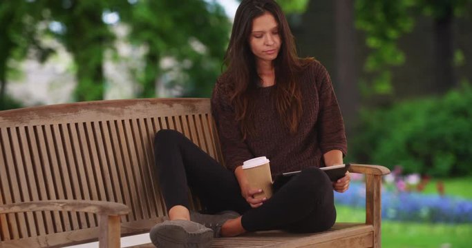 Millennial woman sitting on park bench with flower bed using tablet computer and drinking coffee while visiting Bruges, Tourist sipping drink and using device to plan vacation, 4k
