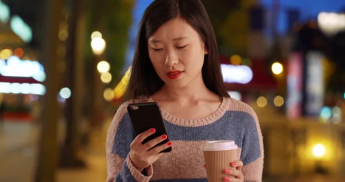 Close up of cute millennial woman reading texts on smartphone and holding cup of coffee in Champs Elysees Paris, Pleasant young Asian woman using cell phone to read text messages in France, 4k