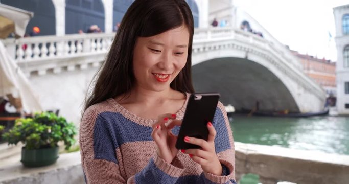 Hip young pretty tourist using smartphone and reading text messages in front of Rialto bridge in Venice Italy, Close up of lovely Asian female millennial sending texts with cellphone from Europe, 4k