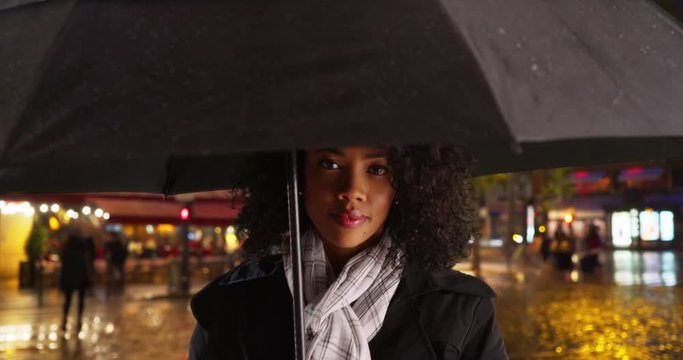 Close up of African American tourist standing under umbrella on rainy night in paris, Millennial woman in black rain coat waiting on street in the rain, 4k