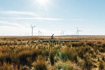 A woman riding a mountain bike in over a wooden track surrounded by wild grass at a wind farm in Scotland