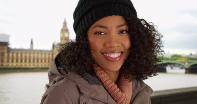 Close up of happy African American woman smiling at camera in front of Big Ben wearing jacket and beanie, Black Millennial tourist on vacation in London standing by the Thames during winter, 4k