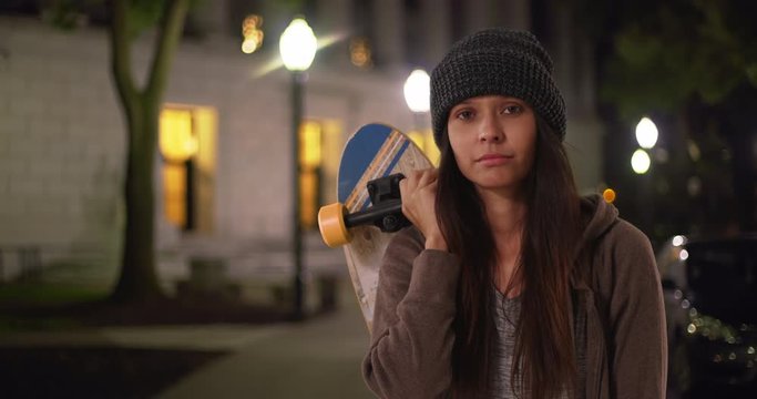Millennial girl holding skateboard over her shoulders looking at camera on street at night, Medium shot portrait of hipster woman with attitude standing in the city, 4k