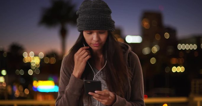 Millennial girl in the city at night text messaging and listening to music on streaming app with mobile phone, Woman with earbuds in texting on cellphone visiting California city, 4k