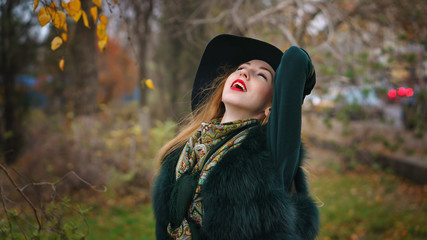 Red-haired girl in autumn park. She is wearing shorts and gloves and wide-brimmed hat. Woman is enjoying good weather.