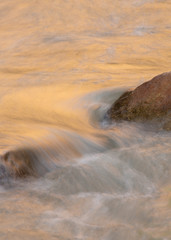 Water flowing over river rocks with gold reflections from the nearby cliffs of Zion National park.  Slow shutter speed used to smooth the water.
