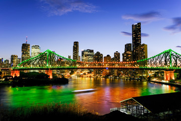View of Brisbane lit up at dusk with a ferry passing under the Story Bridge