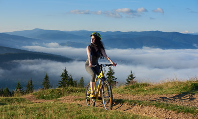 Fototapeta na wymiar Young female cyclist riding on yellow bicycle on a rural trail in the mountains, wearing helmet, enjoying sunny morning. Foggy mountains, forests on the blurred background. Outdoor sport activity