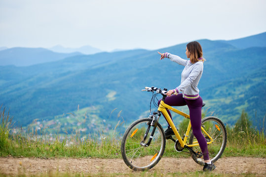 Sporty female cyclist riding on yellow bicycle on a rural trail, pointing at something in the distance. Mountains on the blurred background. Outdoor sport activity. Copy space