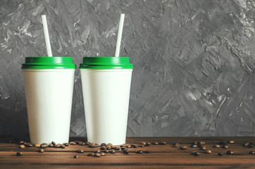 two white plastic coffee cups with a green lid on a wooden table with scattered coffee beans, front view with copy space