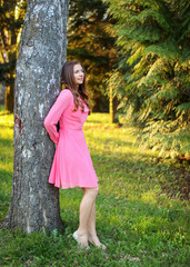 Obraz na płótnie Canvas Young woman wearing pink dress, leaning her back to a tree, with sunset light park in background.