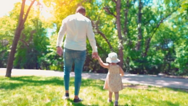 Father with a little daughter walking along the green park holding hands, back view, dad and daughter
