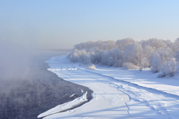 Winter landscape in Siberia. The river does not freeze and above it rises the steam. Trees covered...