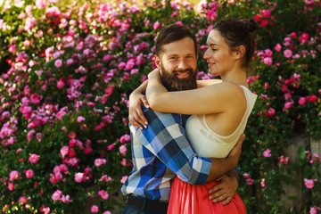 Romantic happy couple in love hugs and look at each other. attractive brunette woman and her handsome bearded boyfriend in park. Bush of roses flowers in the background