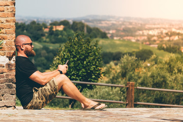 Bearded man sits leaning against a wall of a medieval castle and look on his smartphone instead of...