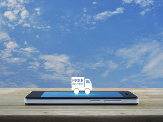 Fototapeta na wymiar Free delivery truck icon on modern smart phone screen on wooden table over blue sky with white clouds, Transportation business concept